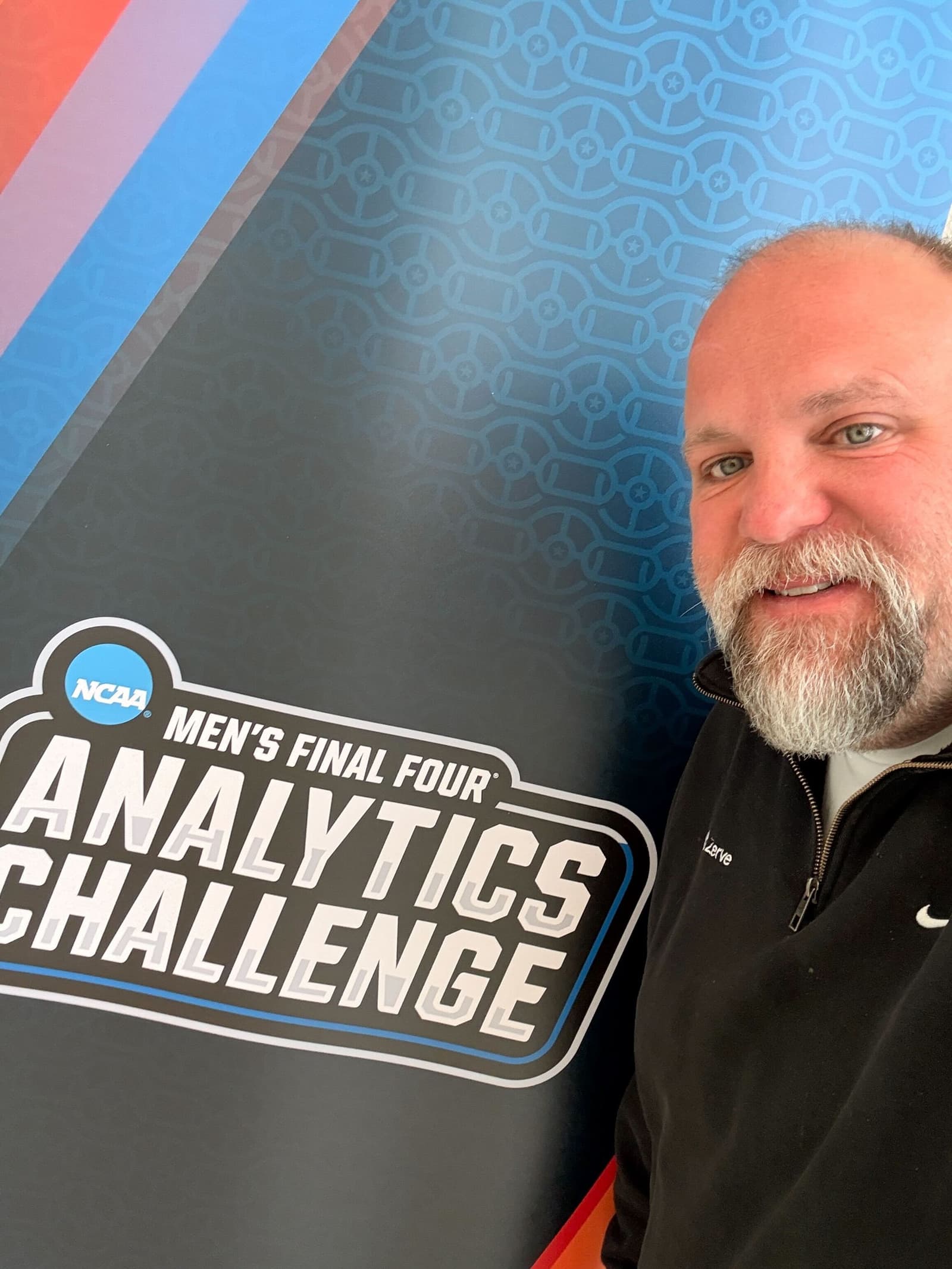 Man smiling near a sign that reads "Men's Final Four Analytics Challenge" with a geometric background.