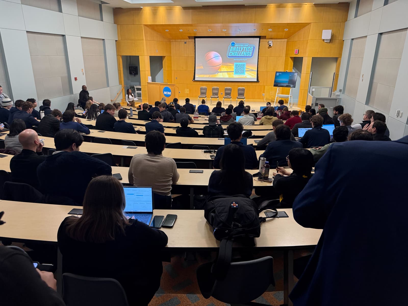 A lecture hall filled with people seated, facing a large screen displaying a basketball and the text "Analytics Challenge" at the front.