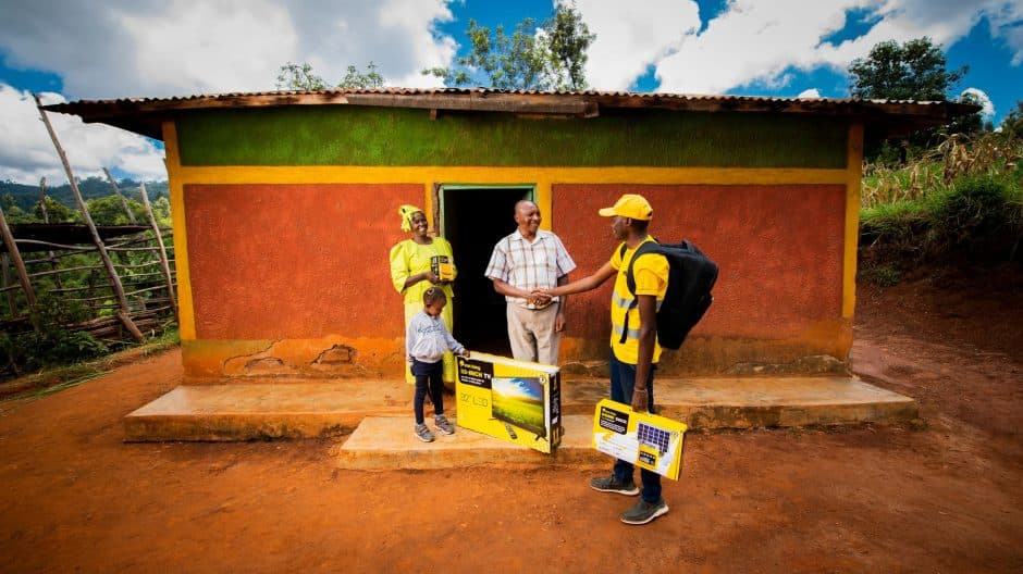 A Sun King representative in a yellow uniform shakes hands with a man outside a brightly painted rural home as he delivers solar-powered products. A woman and child stand nearby holding a solar TV and lighting kit, symbolizing access to renewable energy in rural communities.